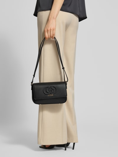 Frauenmodel trägt Handtasche in Leder-Optik Modell 'Isola' von Guess, Schwarz mit Logo-Detail, Magnetverschluss, Hauptfach, Verstellbarer Schulterriemen, Ganzkörperansicht von vorne.