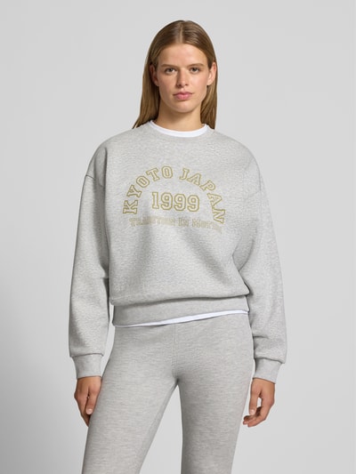 Frauenmodel trägt Sweatshirt aus Baumwoll-Mix Modell 'MERLE' von JIXX in Hellgrau mit Statement-Print, lockerer Passform und gerippten Abschlüssen, Ganzkörperansicht von vorne.