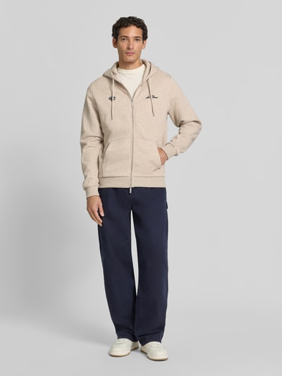 Männermodel trägt Regular Fit Sweatjacke aus reiner Baumwolle mit Logo-Stickerei von Les Deux in Beige, Kängurutasche, Ganzkörperansicht von vorne.