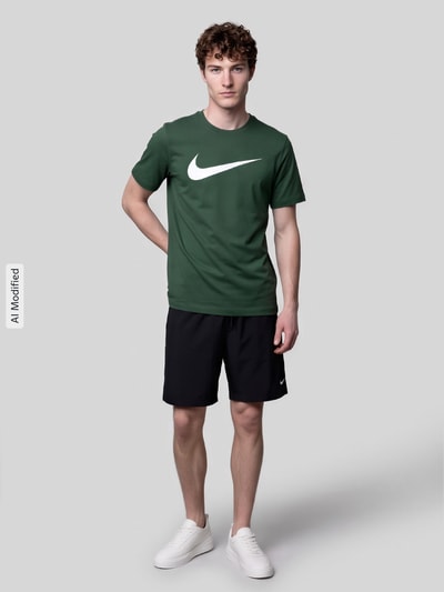 Męski model trägt Regular Fit T-Shirt aus reiner Baumwolle von Nike w kolorze zielonym mit białym logo, czarne spodenki, Ganzkörperansicht von vorne, KI-modifiziertes Bild.