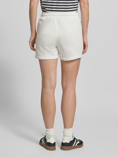 Modelka kobieca ma na sobie Sweatshorts mit Eingrifftaschen und Label-Stitching od Gant, bawełniany mix, krój regular fit, elastyczny pas, kieszenie w szwie, haftowane logo, kolor biały, szerokość w rozmiarze S: 36 cm, długość nogawki wewnętrznej w rozmiarze S: 10 cm, widok całej sylwetki z tyłu.