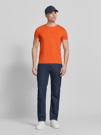 Męski model trägt Slim Fit T-Shirt aus Baumwoll-Mix von Tommy Hilfiger, efekt: jednolity kolor, krój slim fit, detal z logo, okrągły dekolt, krótki rękaw, proste zakończenie rękawów, wykończenie materiału: mieszanka bawełny; ganzkörperansicht von vorne.