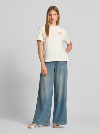 Frauenmodel trägt Relaxed Fit T-Shirt mit Ziersteinbesatz Modell 'Emotional' von GUESS in Weiß, kombiniert mit Jeans, Nahaufnahme Oberkörper. Damen T-Shirt aus reiner Baumwolle mit Logo-Stitching und Ziersteinbesatz, Rückenlänge Größe S: 64 cm.