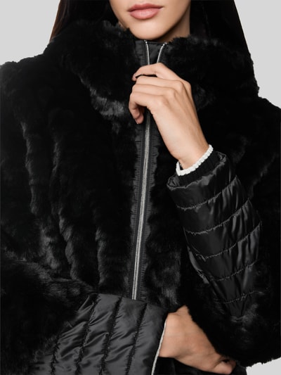 Frauenmodel trägt Mantel aus Kunstfell mit Zweiwege-Reißverschluss von Marc Cain in Schwarz mit Stehkragen, Nahaufnahme Oberkörper. Regular Fit, Rückenlänge 90 cm, Schulterbreite 43 cm, Brustweite 52 cm, Ärmellänge 63 cm.