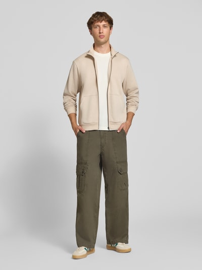 Männermodel trägt Regular Fit Sweatjacke mit Baumwoll-Anteil Modell 'Bradley' von JACK & JONES in Beige, Struktur glatt, Ganzkörperansicht von vorne. Material-Mix, Stehkragen, Reißverschluss, Känguru-Tasche, Rückenlänge 68 cm, Ärmellänge 66 cm.