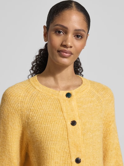 Modelka kobieca ma na sobie Regular Fit Strickjacke aus Woll-Mix mit Alpaka-Anteil Modell 'LULU' od Selected Femme, żółty rippstrick, długie raglanowe rękawy, ściągacze, klasyczny crew neck, jednorzędowe guziki, gładka faktura. Zbliżenie na górną część ciała.