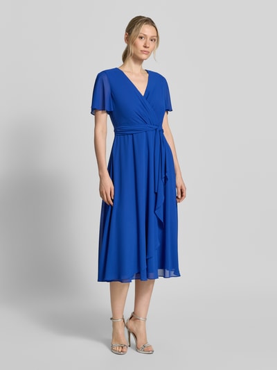 Modelka nosi knielanges Cocktailkleid mit Bindegürtel od Christian Berg Cocktail z szyfonu, taliowany krój, długość do kolan, kopertowy styl, wiązany pasek, długość tyłu w rozmiarze 36: 111 cm, szerokość klatki piersiowej w rozmiarze 36: 38 cm, długość rękawów w rozmiarze 36: 22 cm, intensywny niebieski, gładka tekstura, widok całej sylwetki z przodu.
