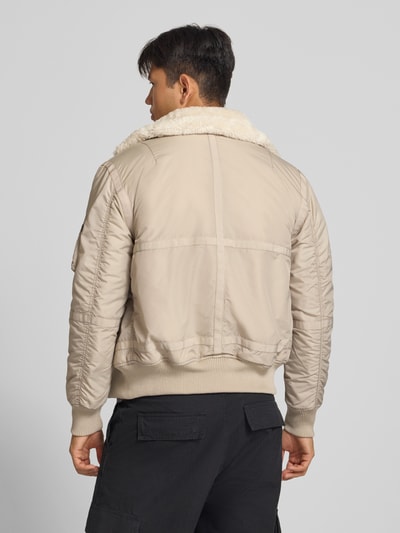 Männermodel trägt Bomberjacke mit Label-Patch und Kentkragen von Alpha Industries in Beige mit Teddyfellkragen, Rückenansicht, Ganzkörperansicht Rückansicht. Regular Fit, Reißverschluss, gerippte Abschlüsse, Reißverschlusstasche am Ärmel, Eingrifftaschen mit Druckknopfverschluss.