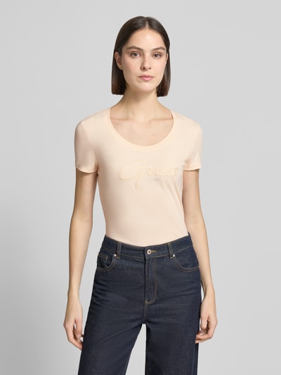 Kobiece model prezentuje damski t-shirt z mieszanki bawełny i elastanu, dopasowany krój, model 'SCRIPT' od Guess, dekolt w łódkę, wyhaftowane logo, długość tyłu w rozmiarze S: 61 cm, jasny kolor, gładka faktura, całkowity widok z przodu