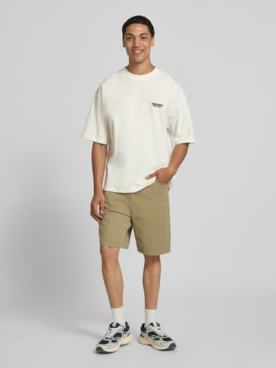 Męski model nosi bermudy z bawełny, krój straight fit, 5 kieszeni, model 'TONY ORIGINAL' od Jack & Jones, kolor beżowy, szerokość w rozmiarze M: 42 cm, długość wewnętrzna nogawki: 20 cm, zapięcie na guzik i zamek błyskawiczny, zdjęcie całej sylwetki z przodu.