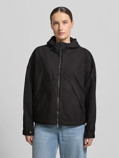 Kobiece model nosi 'Jacke mit Kapuze und Zweiwege-Reißverschluss' od camel active, prosty krój, czarny, wpuszczane kieszenie, regulowane zakończenia rękawów, długość tyłu w rozmiarze 36: 67 cm, szerokość ramion w rozmiarze 36: 44 cm, szerokość w rozmiarze 36: 63 cm, długość rękawów w rozmiarze 36: 62 cm, zamek błyskawiczny dwustronny, kaptur z regulacją, widok całej sylwetki z przodu