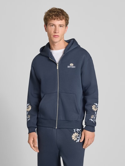 Männermodel trägt Relaxed Fit Sweatjacke mit Kapuze von REVIEW in Dunkelblau, florale Stickerei, Baumwoll-Mix, Ganzkörperansicht von vorne.