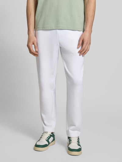 Männermodel trägt weiße Tapered Sweatpants mit Baumwoll-Anteil von Lacoste Sport, glatte Scuba-Optik, Logo-Patch, Ganzkörperansicht von vorne.