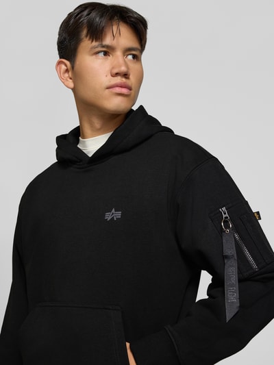 Männermodel trägt Hoodie mit Label-Print von Alpha Industries in Schwarz, Label-Print und Brusttasche, Baumwoll-Mix, Nahaufnahme Oberkörper.