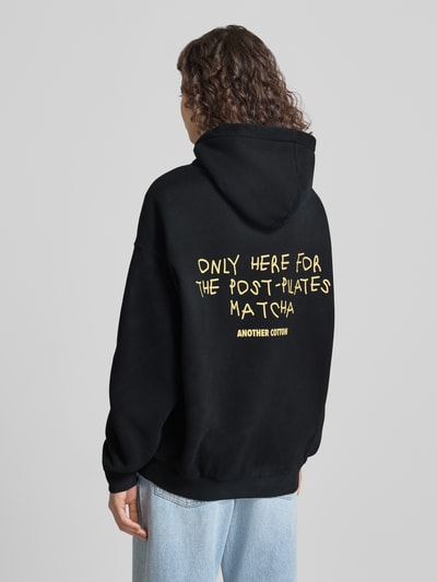 Männermodel trägt czarny Hoodie mit Logo-Stitching od ANOTHER COTTON LAB, mieszanka bawełny, luźny krój, kaptur, kieszeń kangurka, Ganzkörperansicht Rückansicht.