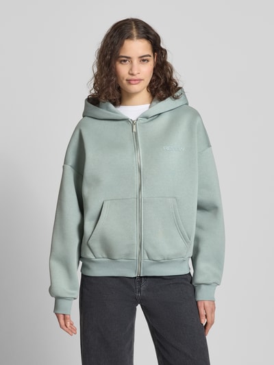 Frauenmodel trägt Sweatjacke mit Logo und Känguru-Tasche von Review in Mintgrün, Relaxed Fit, Baumwoll-Mix, Ganzkörperansicht von vorne.