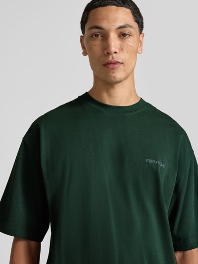 Męski model nosi Oversized T-Shirt aus reiner Baumwolle od REVIEW, zielony, nadruk z logo, okrągły dekolt, bawełna, krój oversized, długość tyłu w rozmiarze M: 66 cm, zbliżenie na górną część ciała.