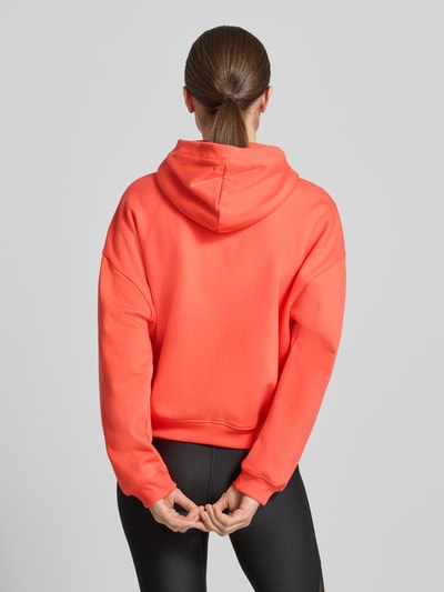 Kobiece model prezentuje Hoodie mit Reißverschluss und Label-Stitching od Under Armour z bawełnianej mieszanki, krój regular fit, pomarańczowy kolor, zdobienie label-stitching, kieszeń kangurka, widok całej sylwetki z tyłu.