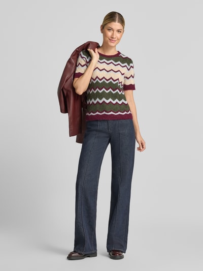 Frauenmodel trägt Strickshirt mit Rundhalsausschnitt von miss goodlife in taillierter Passform, mehrfarbiges Zackenmuster, gerippte Abschlüsse, kurzärmeliges Design, dazu dunkle Jeans und Jacke in der Hand, Ganzkörperansicht von vorne.
