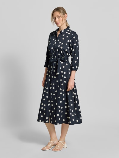 Modelka nosi Regular Fit Blusenkleid aus Baumwoll-Mix od s.Oliver BLACK LABEL, ciemnoniebieska tkanina z białymi grochami, długość midi, pasek w talii, długie bufiaste rękawy, mankiety, kieszenie boczne, widok całej sylwetki z przodu.