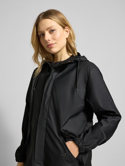 Modelka kobieca nosi Regular Fit Parker z kapturem Model 'Fishtail' od Rains, czarny kolor, z kieszeniami z patką, elastyczne zakończenia rękawów, krój regular fit, długość tyłu w rozmiarze S: 104 cm, szerokość ramion: 45 cm, szerokość w rozmiarze S: 55 cm, długość rękawów: 65 cm, półzbliżenie przód.
