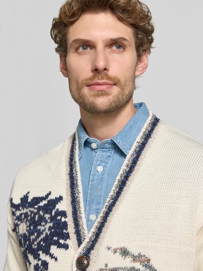 Männermodel trägt Strickjacke mit V-Ausschnitt Modell 'INTARSIA' von Scotch & Soda, cremefarben, Allover-Look, Nahaufnahme Oberkörper. Herrenkontext: Baumwoll-Mix mit Woll-Anteil, Regular Fit, Knopfleiste, Rückenlänge 71 cm, Ärmellänge 60 cm.