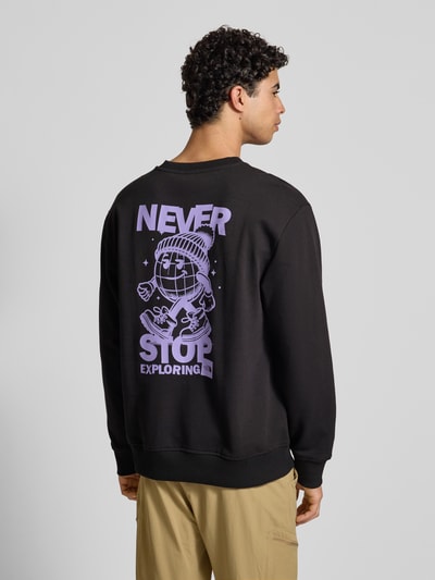 Męski model nosi bluzę Sweatshirt mit Label-Print od The North Face z czarnej bawełny, krój relaxed fit, nadruk z logo, prążkowane wykończenia, okrągły dekolt, długość tyłu 70 cm, długość rękawa 65 cm, całkowita sylwetka z tyłu.