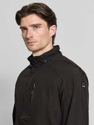 Mężczyzna nosi Jacke mit Napoleontasche Modell 'KOS' od Killtec, czarny kolor, detal napoleońskiej kieszeni, label na ramieniu, tkanina o gładkiej fakturze, prosta linia ramion, regular fit; zbliżenie na górną część ciała, ujęcie z przodu. Szerokość w rozmiarze M: 60 cm, długość tyłu: 73 cm, szerokość ramion: 46 cm, długość rękawa: 71 cm.
