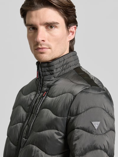 Männermodel trägt Steppjacke mit Reißverschluss von Guess in Anthrazit, Stehkragen, glatte und gesteppte Textur, Nahaufnahme Oberkörper. Kontext: Regular Fit, Reißverschlusstaschen, gerippte Abschlüsse, Maße Größe M 71/50/60/69 cm.