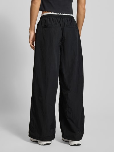 Kobiece model prezentuje Spodnie typu track pants Regular Fit Trackpants mit Kontraststreifen von Tommy Jeans, efekt: jednolity kolor, krój regular fit, aplikacja: paski w kontrastowym kolorze, pas z logo, zapięcie: tunel, ściągacz: elastyczny, podwójny pas, kieszenie zapinane na zamek błyskawiczny po bokach, kieszeń z tyłu, widok całej sylwetki z tyłu.
