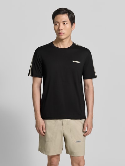 Męski model nosi Regular Fit T-Shirt mit Logo-Patch od EA7 Emporio Armani z bawełny, okrągły dekolt, czarny, długość tyłu w rozmiarze M: 52 cm, widok z przodu, ujęcie całej sylwetki.