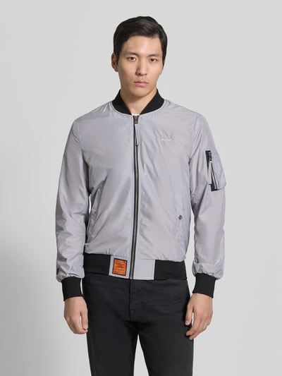 Männermodel trägt silbergraue Bomberjacke mit Label-Patch und Eingrifftaschen von Original Bombers, Stoff leicht glänzend, Ganzkörperansicht von vorne, Gerader-Schnitt, Reißverschluss, Reißverschlusstasche am Ärmel, Druckknopfverschluss, Gerippte-Abschlüsse, Rückenlänge bei Größe M: 67 cm, Schulterbreite: 43 cm, Brustweite: 53 cm, Ärmellänge: 68 cm.