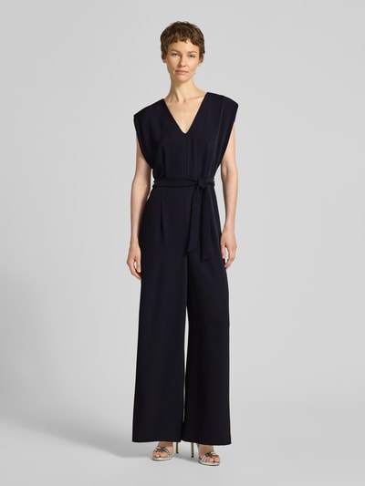 Frauenmodel trägt Jumpsuit mit V-Ausschnitt und Bindegürtel von comma, dunkle Farbe, glatte Textur, Regular Fit, Ganzkörperansicht von vorne.