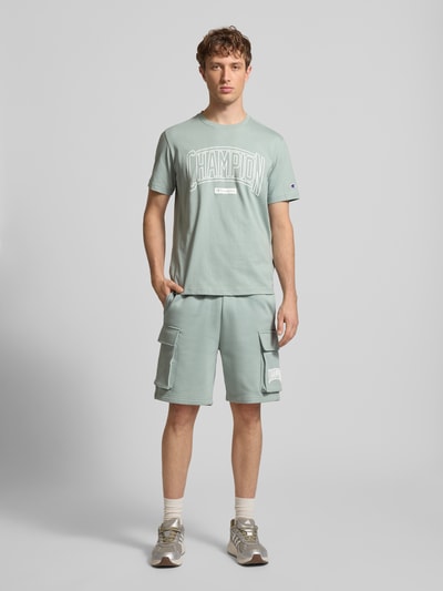 Männermodel trägt T-Shirt mit Logo und Rundhalsausschnitt von CHAMPION aus reiner Baumwolle im Regular-Fit mit Label-Stitching, Ganzkörperansicht von vorne.