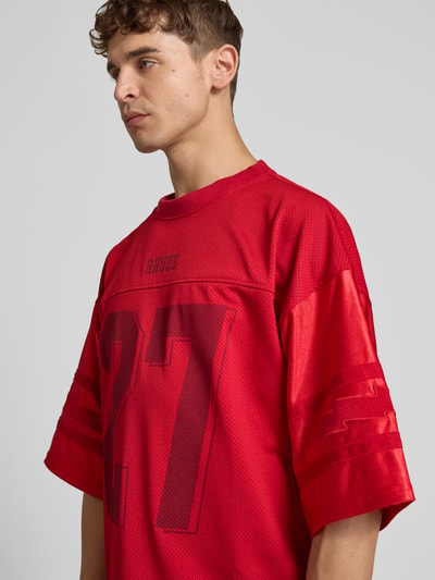 Männermodel trägt Relaxed Fit T-Shirt mit 3/4-Ärmel von RHUDE in Rot mit Statement-Print und Satin-Mesh, Ganzkörperansicht von vorne. Herren T-Shirt mit Rundhalsausschnitt, Rückenlänge 69 cm bei Größe M, Label-Detail.