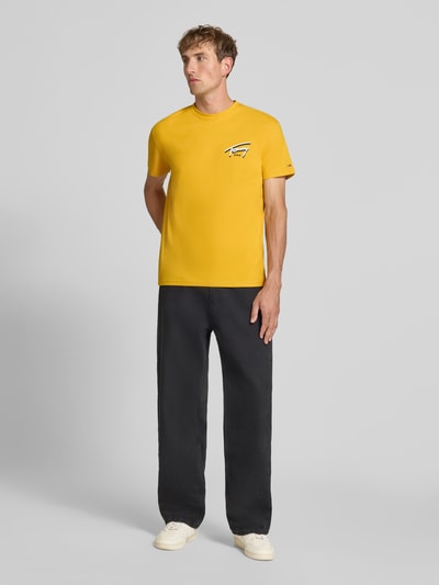 Männermodel trägt Regular Fit T-Shirt aus reiner Baumwolle von Tommy Jeans in Gelb zu dunkler Hose, Ganzkörperansicht von vorne.