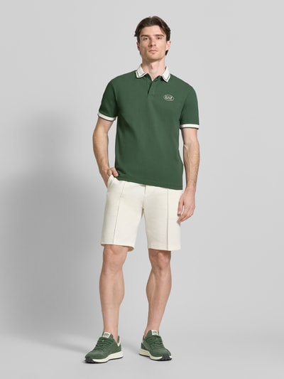 Mężczyzna prezentuje Regular Fit Poloshirt mit Logo-Print od EA7 Emporio Armani, elastyczna bawełna, polokrótki rękaw, krój regular fit, długość tyłu w rozmiarze M 68 cm, widok całej sylwetki z przodu.