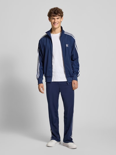 Männermodel trägt Trainingshose mit Kontraststreifen und Reißverschlusstaschen von adidas Originals in Dunkelblau, Regular Fit, Modell FIREBIRD, Elastikbund, Label-Stitching, Ganzkörperansicht von vorne.