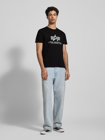 Męski model nosi T-shirt mit Label-Print od Alpha Industries, czarny, bawełna, prosty krój, okrągły dekolt, długość tyłu w rozmiarze L: 70 cm, całopostaciowy widok z przodu.