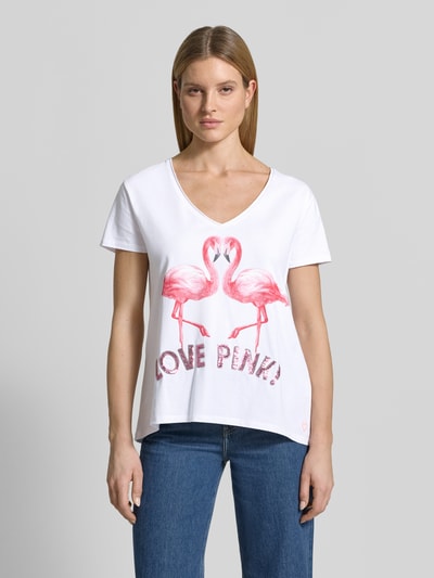 Kobiece modelka nosi damski t-shirt z mieszanki bawełny i elastanu, krój regular fit, model 'Flamingo Love Me' od miss goodlife, dekolt w serek, nadruk z motywem flaminga, długość tyłu w rozmiarze S: 66 cm, jasne kolory, przód zdobi nadruk, widoczne miękkie tekstury, widok całej sylwetki z przodu.