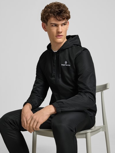 Männermodel trägt Regular Fit Tracksuit mit Logo-Stitching Modell 'Carson' von SERGIO TACCHINI in Schwarz, Nahaufnahme Oberkörper. Herren, Kapuze, Tunnelzug, Reißverschluss, elastische Abschlüsse, Eingrifftaschen, Bundweite 38 cm, Rückenlänge 67 cm, Schulterbreite 43 cm, Ärmellänge 70 cm, Innenbeinlänge 76 cm.