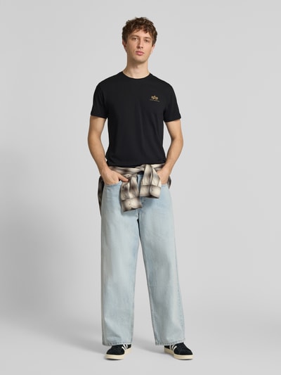 Männermodel trägt T-Shirt aus Baumwolle von Alpha Industries in Schwarz mit Label-Details, Gerader-Schnitt, Rundhalsausschnitt, Ganzkörperansicht von vorne.