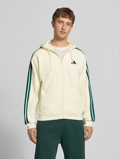 Männermodel trägt Regular Fit Sweatjacke aus Baumwoll-Mix von adidas, cremefarben mit grünen Streifen, adidas Sportswear, Ganzkörperansicht von vorne.