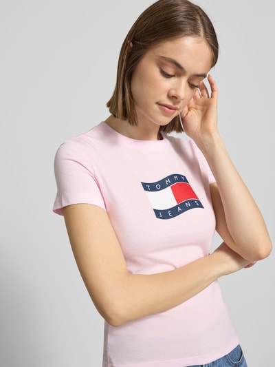 Frauenmodel trägt Slim Fit T-Shirt aus reiner Baumwolle von Tommy Jeans mit Optik: Unifarben, Passform: Slim-Fit, Label-Print, Rundhals-Ausschnitt, Ärmelform: Kurzarm, Materialverarbeitung: Jersey, Farbe Hellrosa, Nahaufnahme Oberkörper.