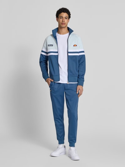 Męski model nosi bluza rozpinana z mieszanki bawełny model 'RIMINI' od Ellesse, niebieskie i białe kolory, prosta sylwetka, stójka, zamek błyskawiczny, detale z logo, elastyczne wykończenia, długość tyłu w rozmiarze M: 71 cm, długość rękawów: 43 cm, widok całej sylwetki z przodu.