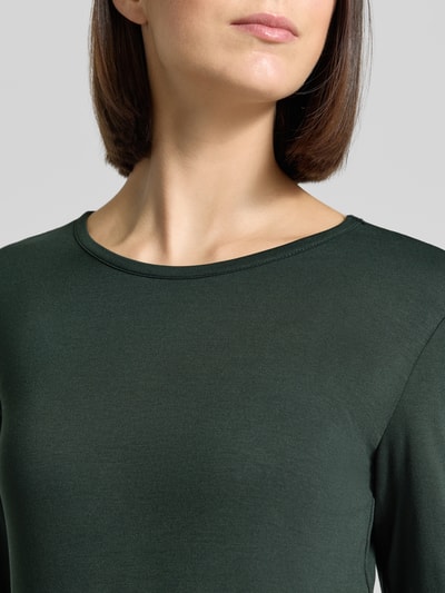 Kobieta nosi bluzkę z długim rękawem Longsleeve aus Viskose-Elasthan-Mix Modell 'Liana' od FREE/QUENT, krój slim fit, okrągły dekolt, mieszanka wiskozy i elastanu, długość tyłu w rozmiarze S: 59 cm, ciemnozielona, gładka faktura, zbliżenie na górną część ciała