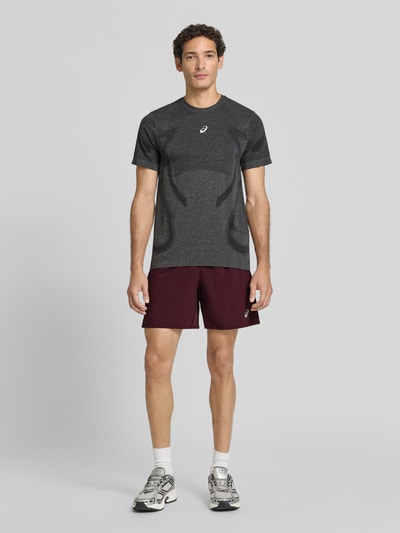 Männermodel trägt Shorts mit elastischem Bund und Logo von ASICS in Dunkelrot, kombiniert mit Innenhose, Regular Fit, Ganzkörperansicht von vorne.