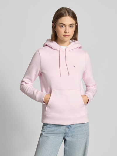 Kobieta modelka nosi Regular Fit Hoodie aus Baumwoll-Mix od Tommy Jeans, jasnoróżowy, jednolity, kieszeń, długi rękaw, widok z przodu do pasa.