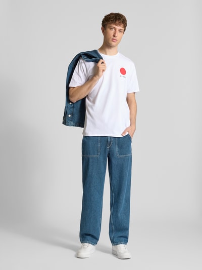 Męski model nosi białe T-shirt aus reiner Baumwolle Modell 'JAPANESE SUN' od Edwin, prosty krój, okrągły dekolt, jeansy, sneakersy, widok całej sylwetki z przodu.