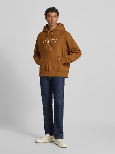 Männermodel trägt Hoodie mit Label-Print und Känguru-Tasche van CK Calvin Klein in bruin, mix van katoen en elastaan, relaxed fit, ganzkörperansicht von vorne, ruglengte bij maat M 69 cm, mouwlengte 60 cm.